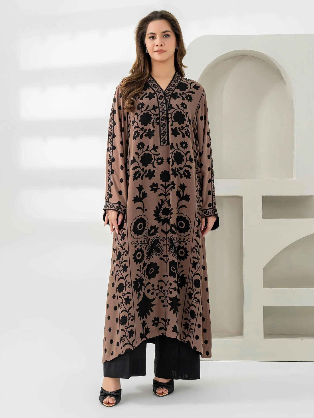 A woman wearing a long, loose-fitting brown and black printed kurta paired with black trousers stands against a minimal white background.