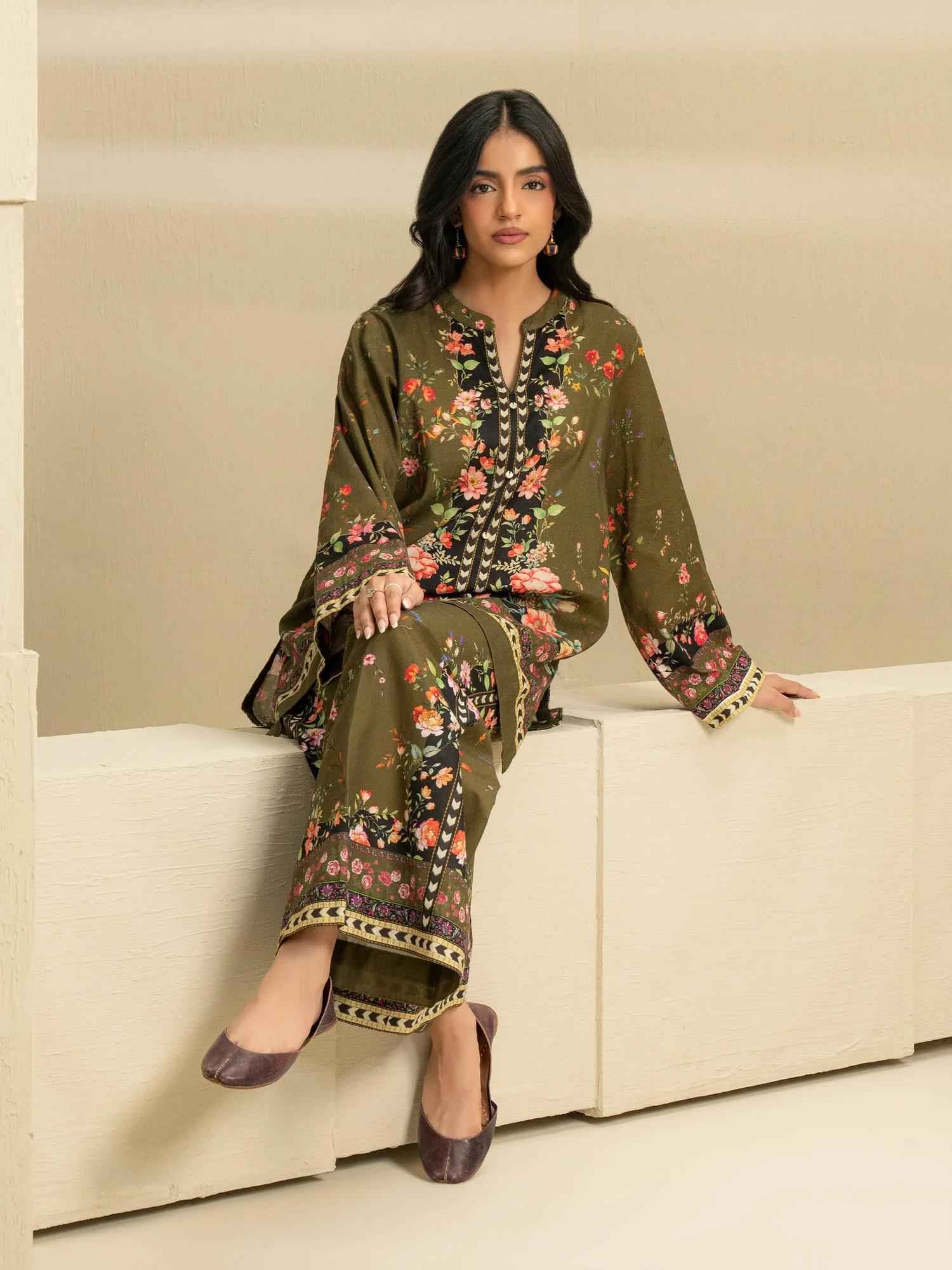 A woman wearing an olive-green embroidered Pakistani suit sits on a white step, posing indoors against a neutral background.