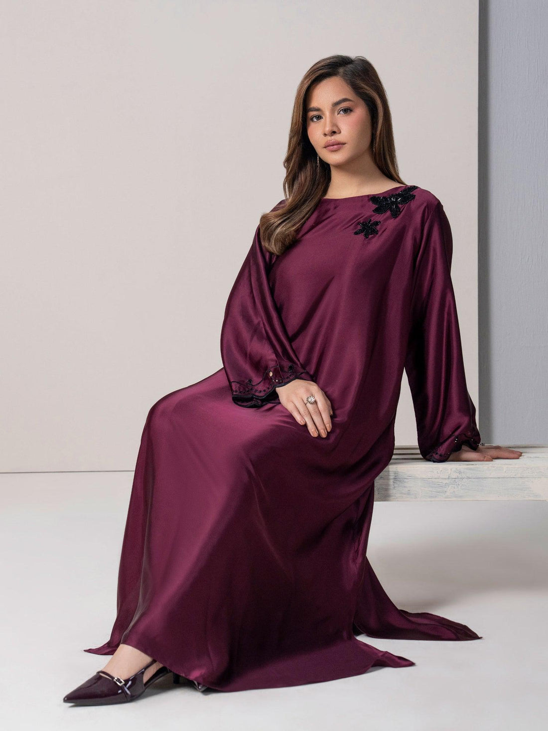 A woman wearing a long maroon Pakistani dress, sitting gracefully against a minimal studio backdrop