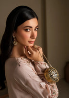 A woman with long black hair is shown in a close-up portrait, wearing a pale blush pink embroidered top and holding a round gold embellished clutch bag with a chain strap.