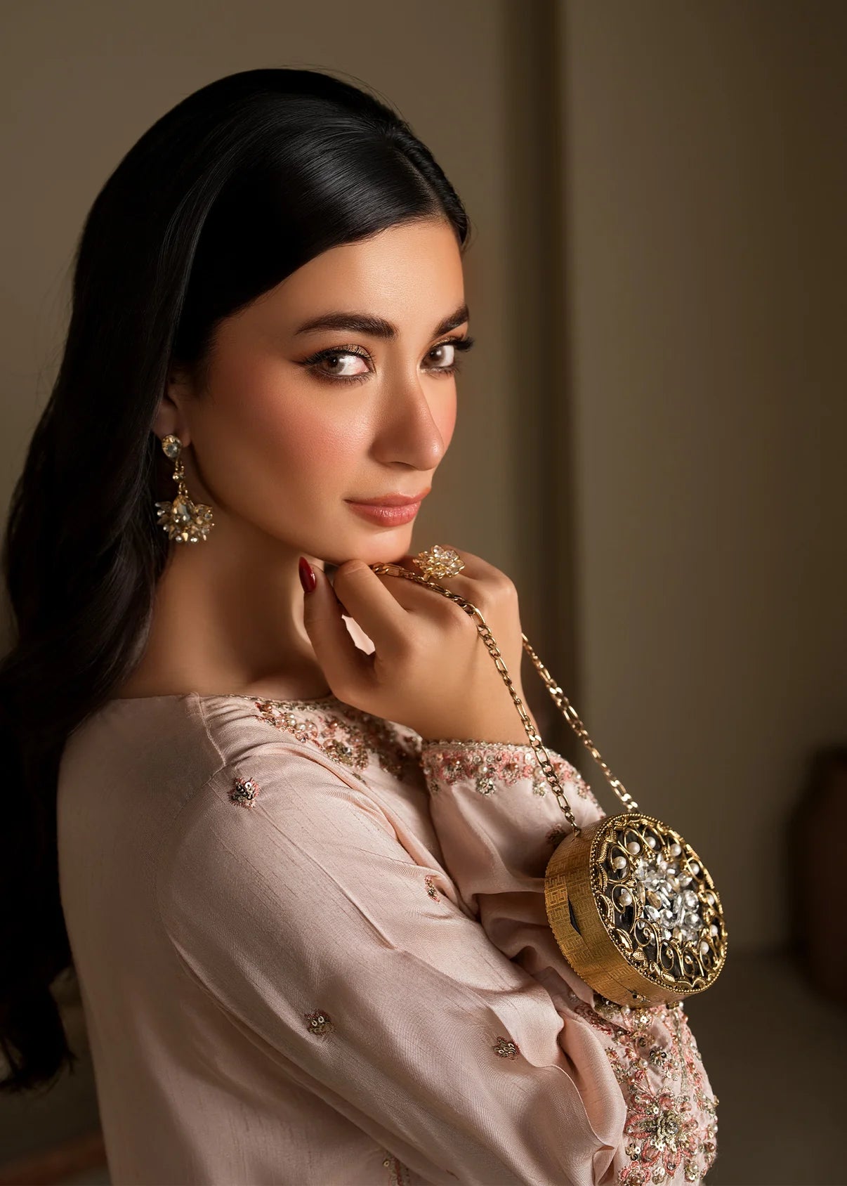 A woman with long black hair is shown in a close-up portrait, wearing a pale blush pink embroidered top and holding a round gold embellished clutch bag with a chain strap.