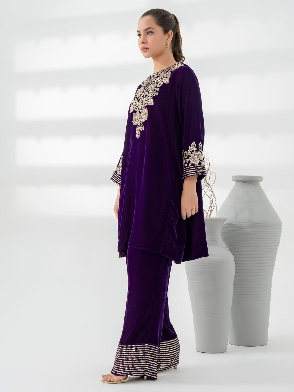 Woman wearing a purple outfit with intricate gold embroidery standing next to white vases on a light gray background