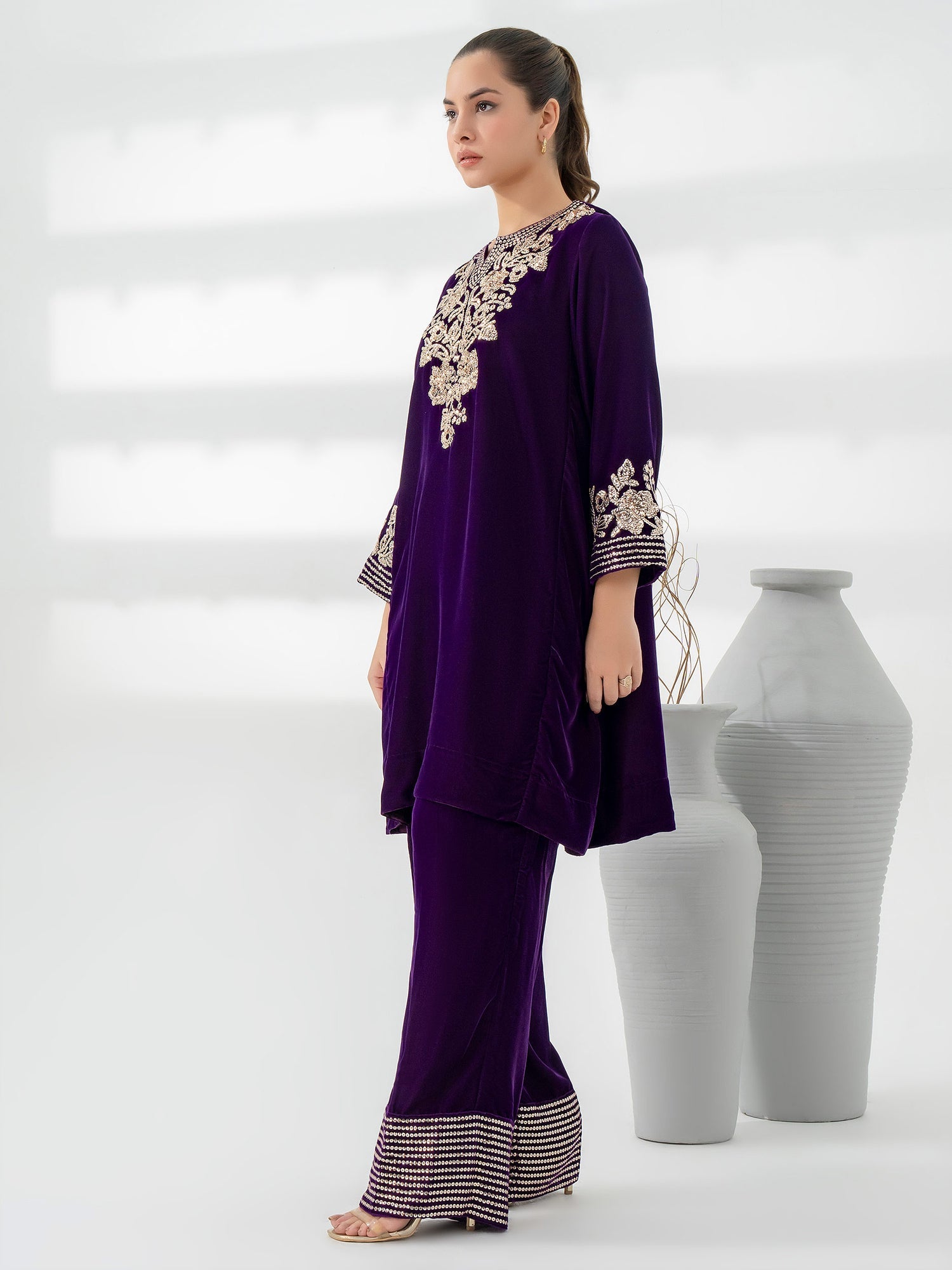 Woman wearing a purple outfit with intricate gold embroidery standing next to white vases on a light gray background
