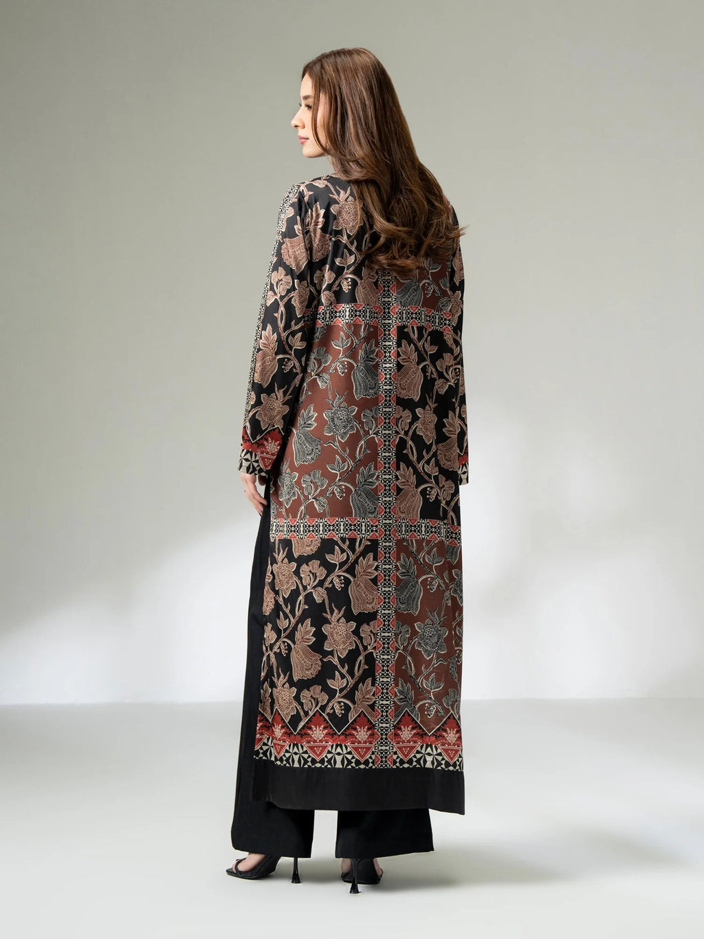 Woman wearing a long printed silk shirt in black and earthy tones with floral patterns and button detailing, paired with black trousers