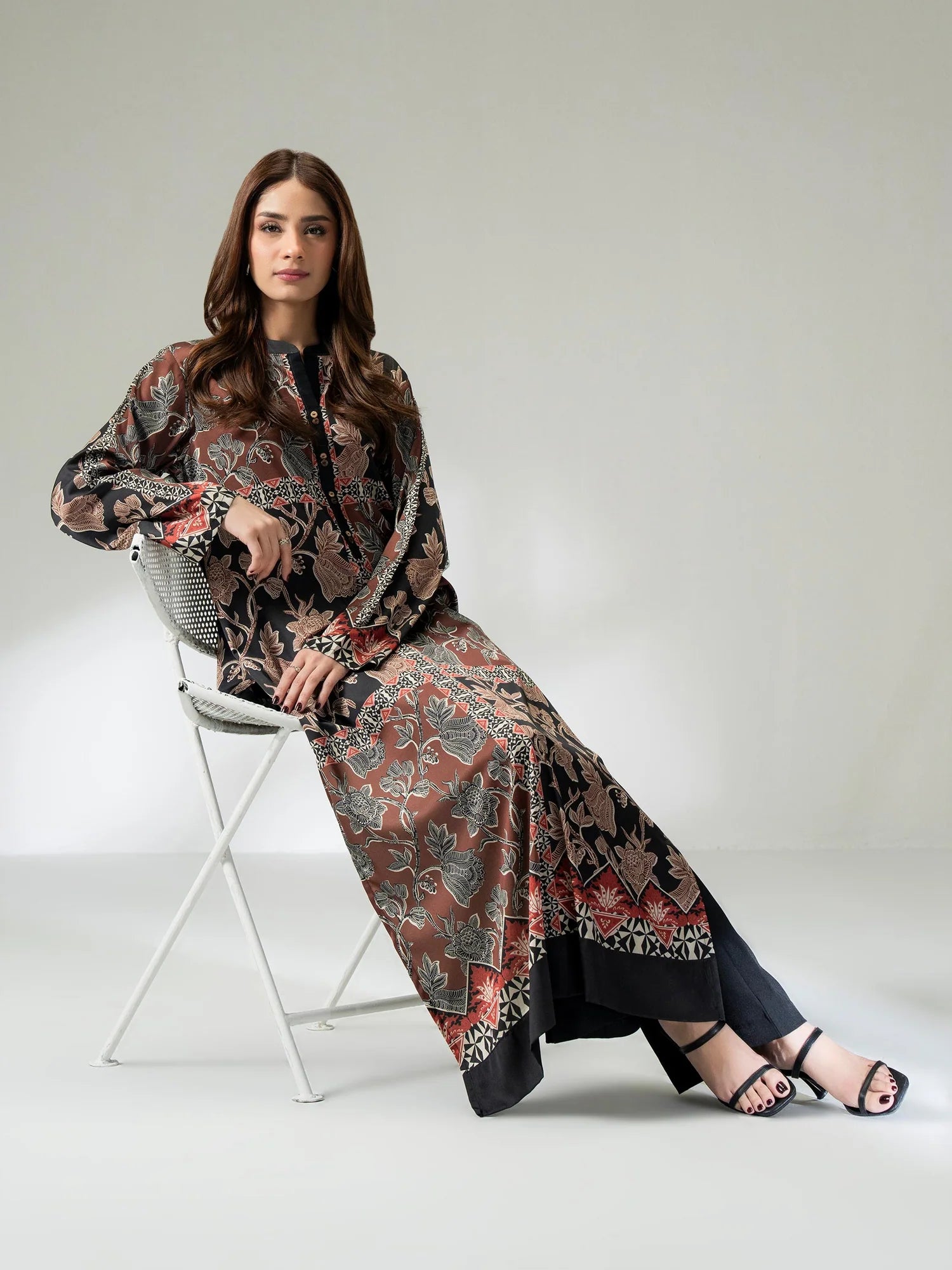 Woman wearing a long printed silk shirt in black and earthy tones with floral patterns and button detailing, paired with black trousers