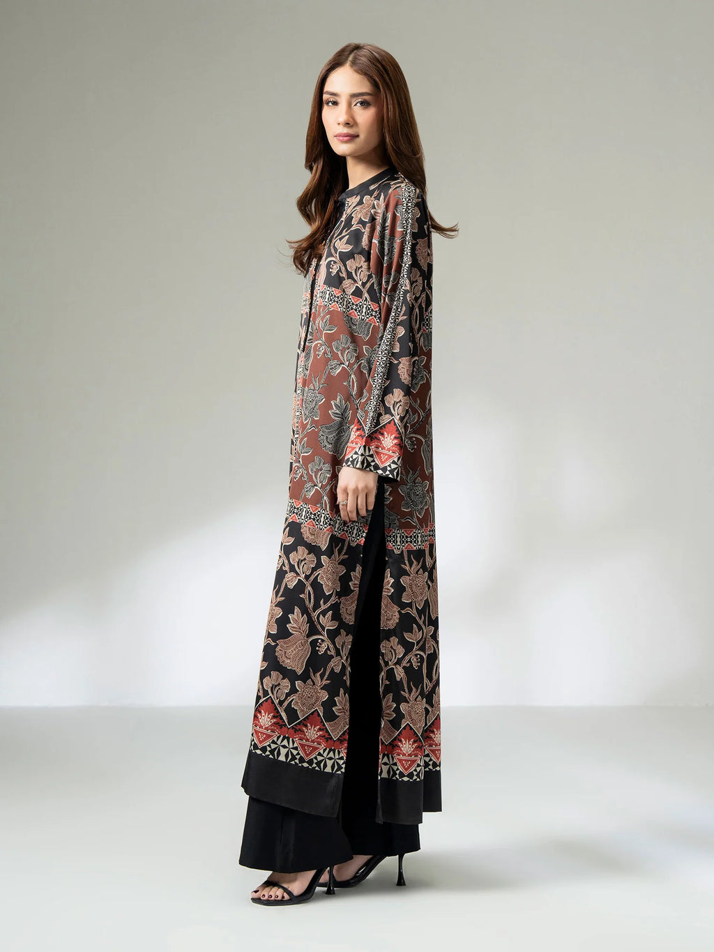 Woman wearing a long printed silk shirt in black and earthy tones with floral patterns and button detailing, paired with black trousers