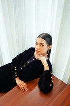 Woman in a black outfit with gold buttons leaning on a wooden surface against white curtains.