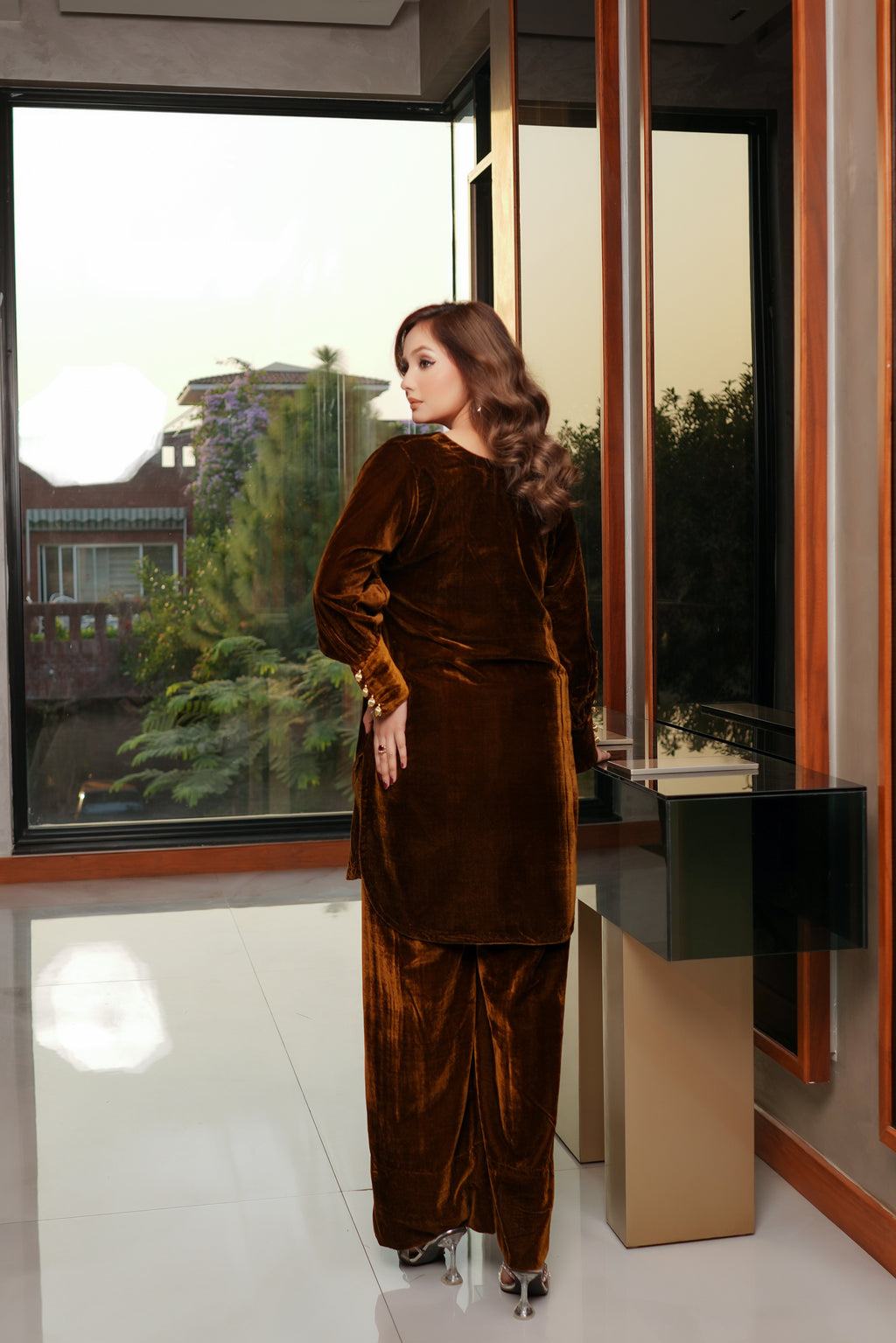 Woman in a brown velvet outfit standing in a modern interior setting with large windows.
