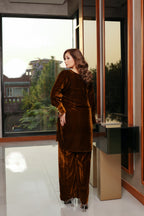 Woman in a brown velvet outfit standing in a modern interior setting with large windows.